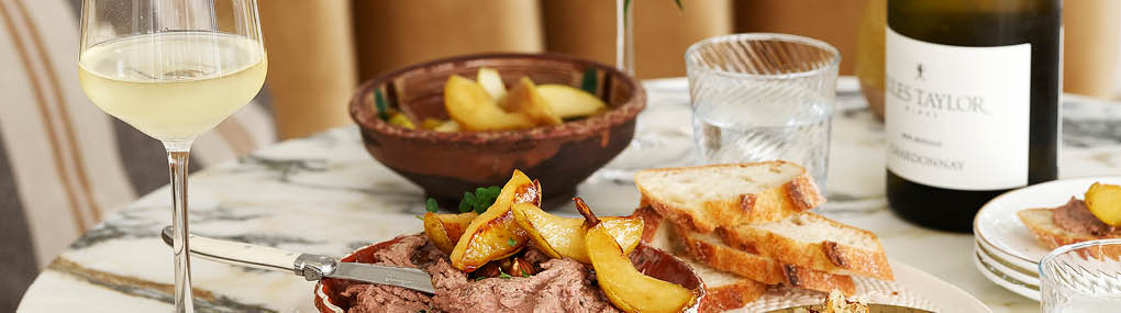 A plate of pate and grilled pears on a marble table, wiht a glass of white wine to the left, and a bottle of Jules Taylor Chardonnay blurred to the right.