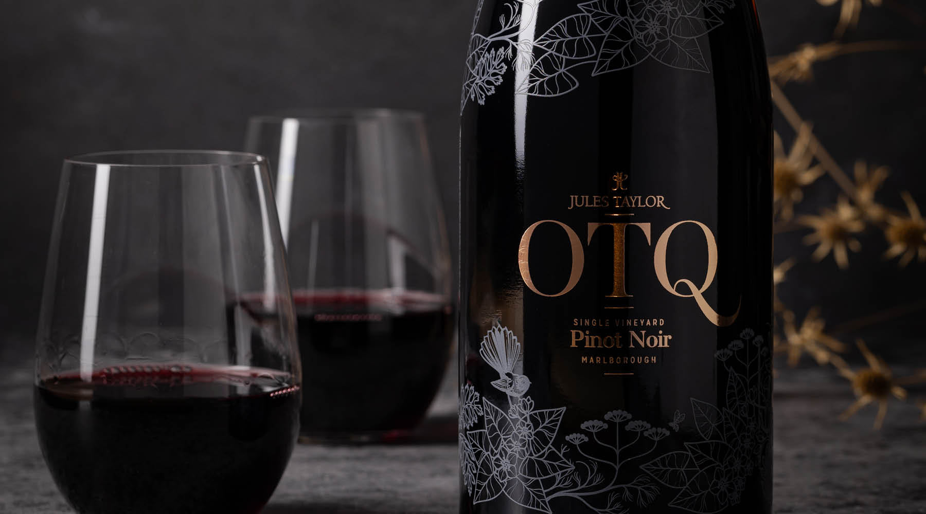 A bottle of Jules Taylor On The Quiet Pinot Noir red wine stands next to two stemmed glasses filled with red wine on a dark tabletop against a dark gray background.