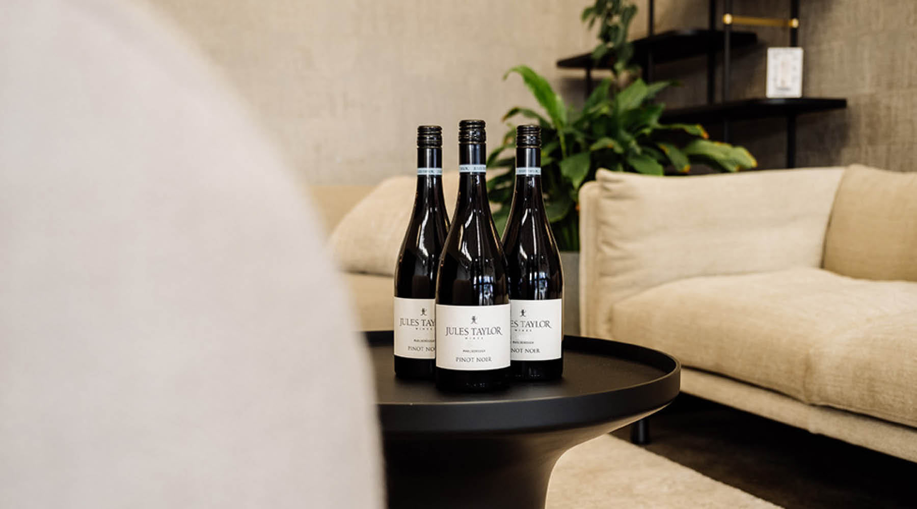 Three bottles of Jules Taylor Pinot Noir arranged on a round black side table in a modern living room setting with a beige sofa and a plant in the background.