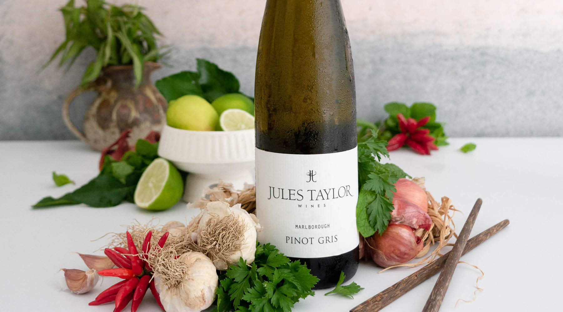 A bottle of Jules Taylor Marlborough Pinot Gris wine stands on a white surface surrounded by fresh ingredients including garlic, parsley, red chilis, limes, shallots, and cinnamon sticks.