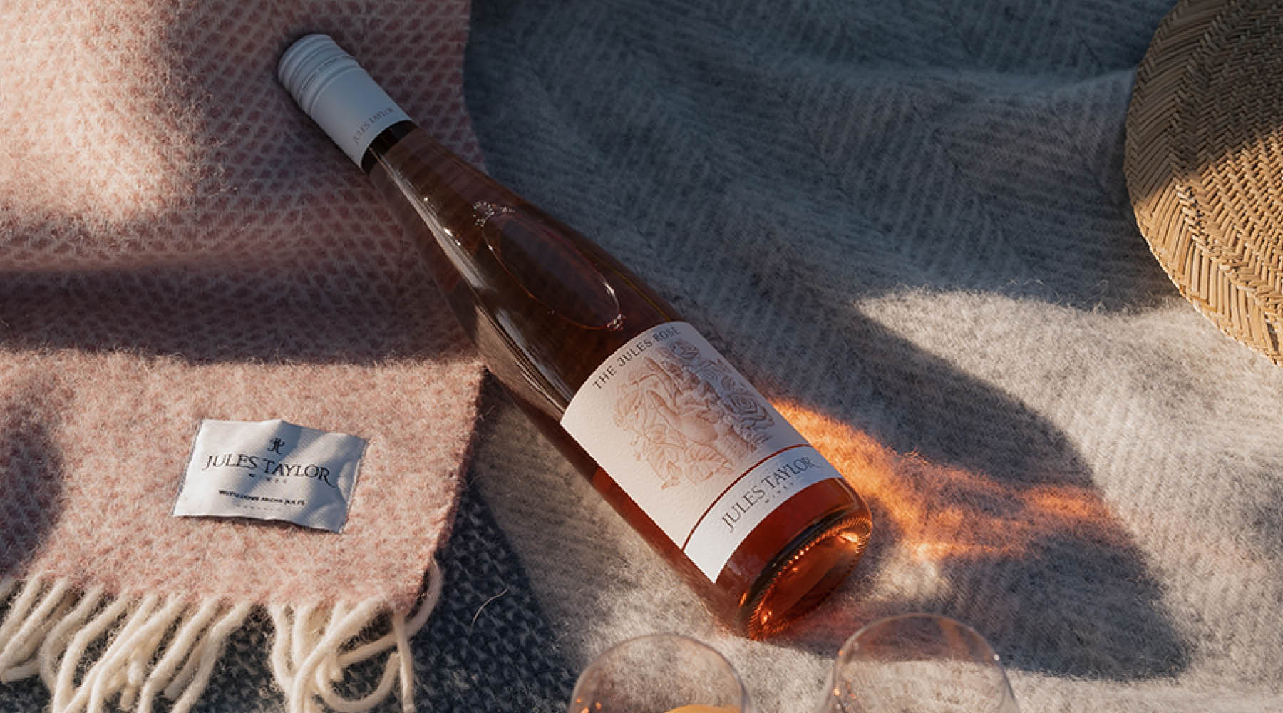 A bottle of Rosé wine by Jules Taylor lies diagonally on a textured pink blanket with fringe, alongside a small branded tag, under warm sunlight and shadow.