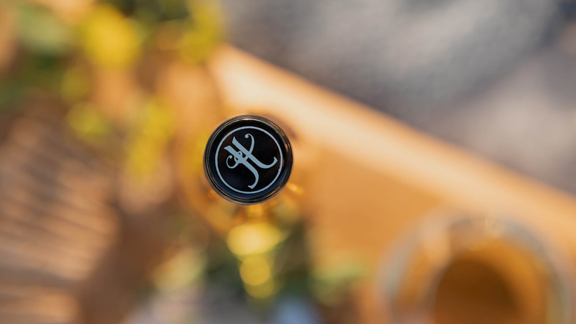 Looking down on a bottle of Jules Taylor Late Harvest Sauvignon Blanc with the Jules Taylor monogram close up and in focus, and a blurred background of warm wood and foliage.