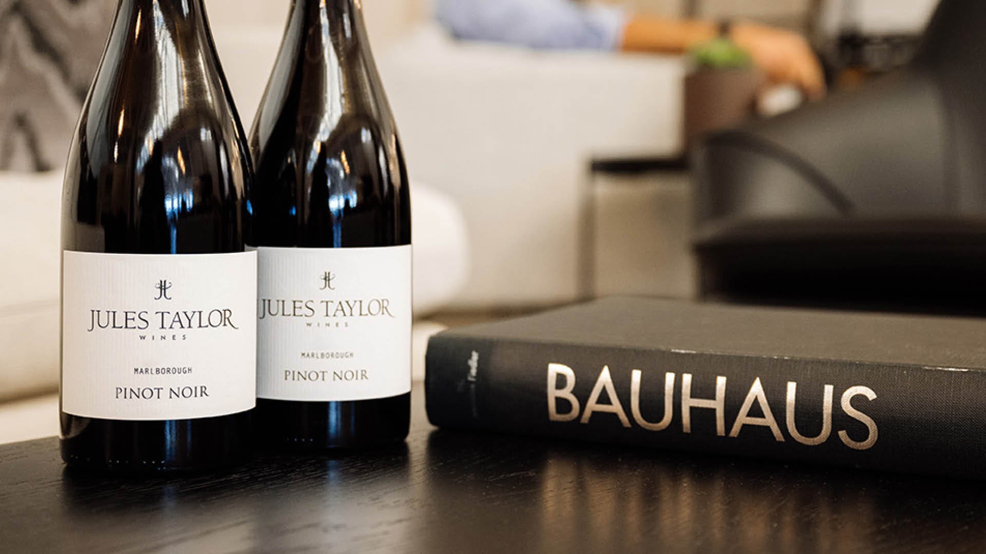 Two bottles of Jules Taylor Pinot Noir stand on a dark table next to a black Bauhaus book, with a softly blurred indoor background.
