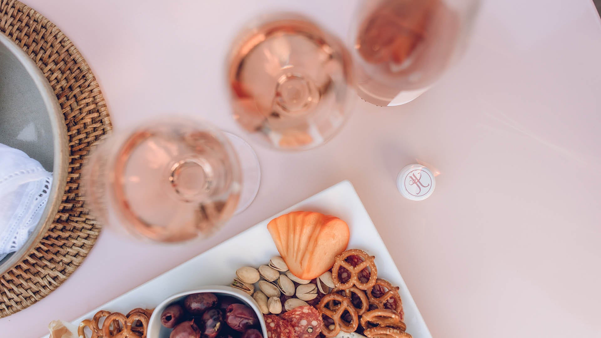 An overhead flat-lay of three glasses of rosé wine accompanied by a charcuterie board with crackers, nuts, and olives, on a light pink surface.