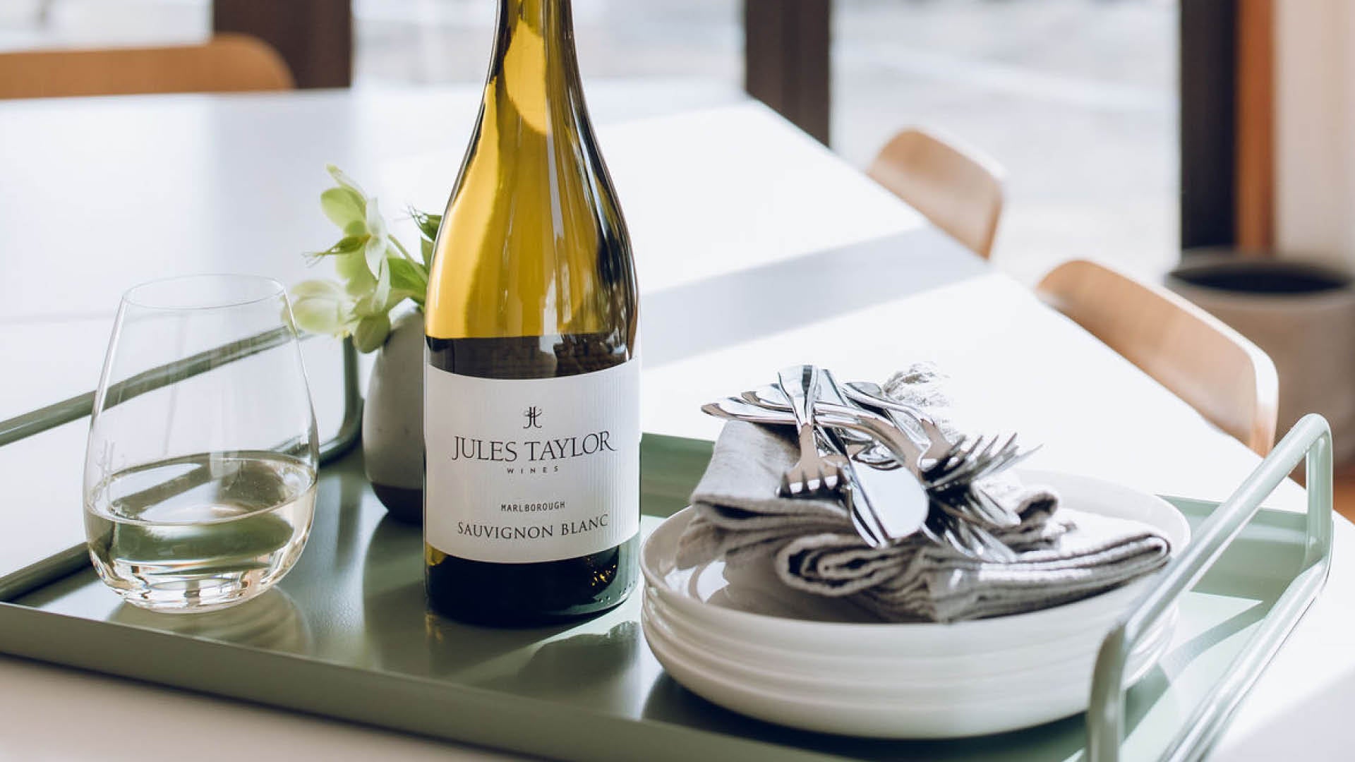 A bottle of Jules Taylor Marlborough Sauvignon Blanc wine, styled on a green tray with a glass, white plates, and silver cutlery on a dining table.