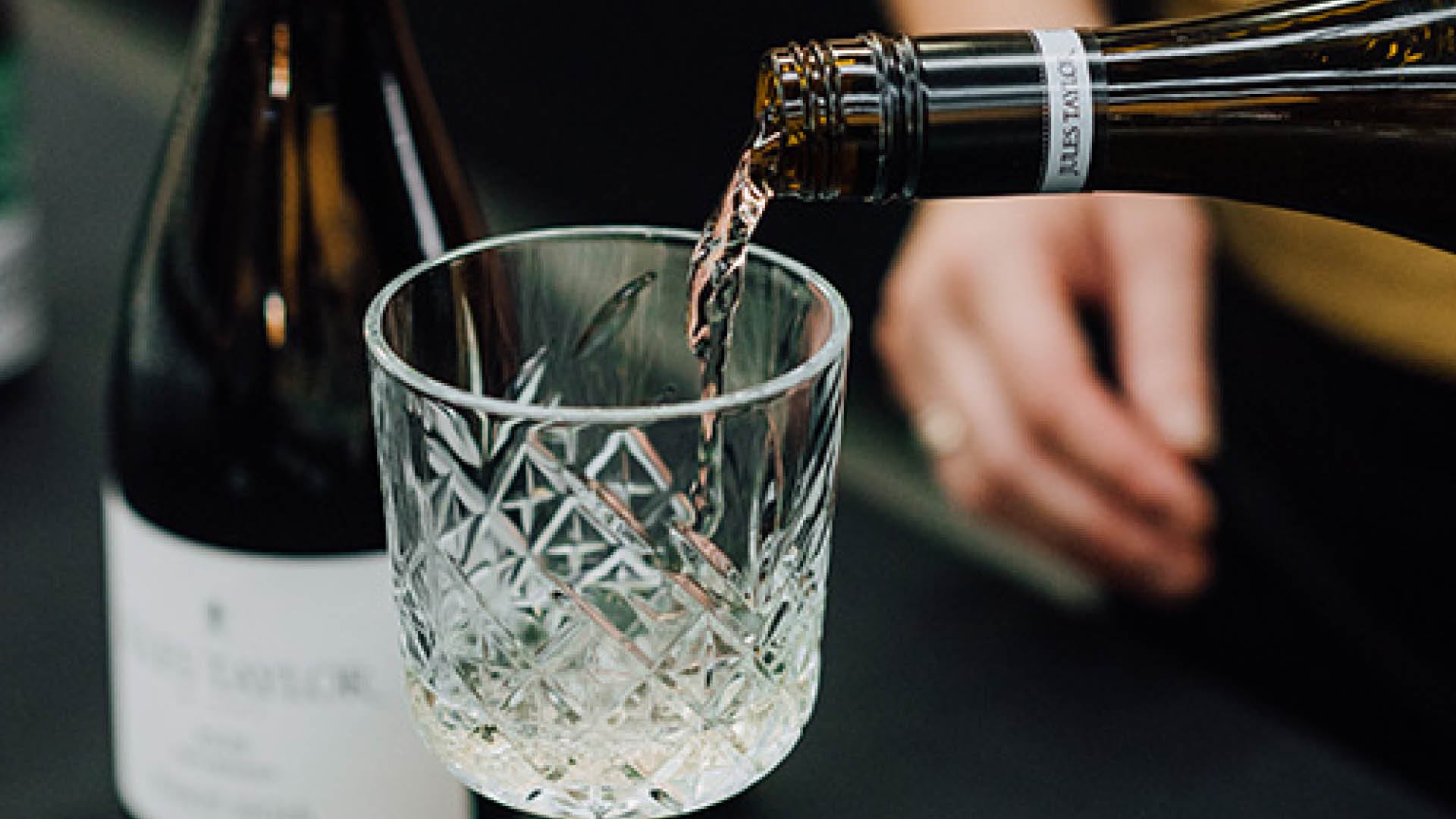 A close-up of a hand pouring Jules Taylor Sauvignon Blanc white wine from a bottle into a clear cut-crystal tumbler, with another wine bottle blurred in the background.
