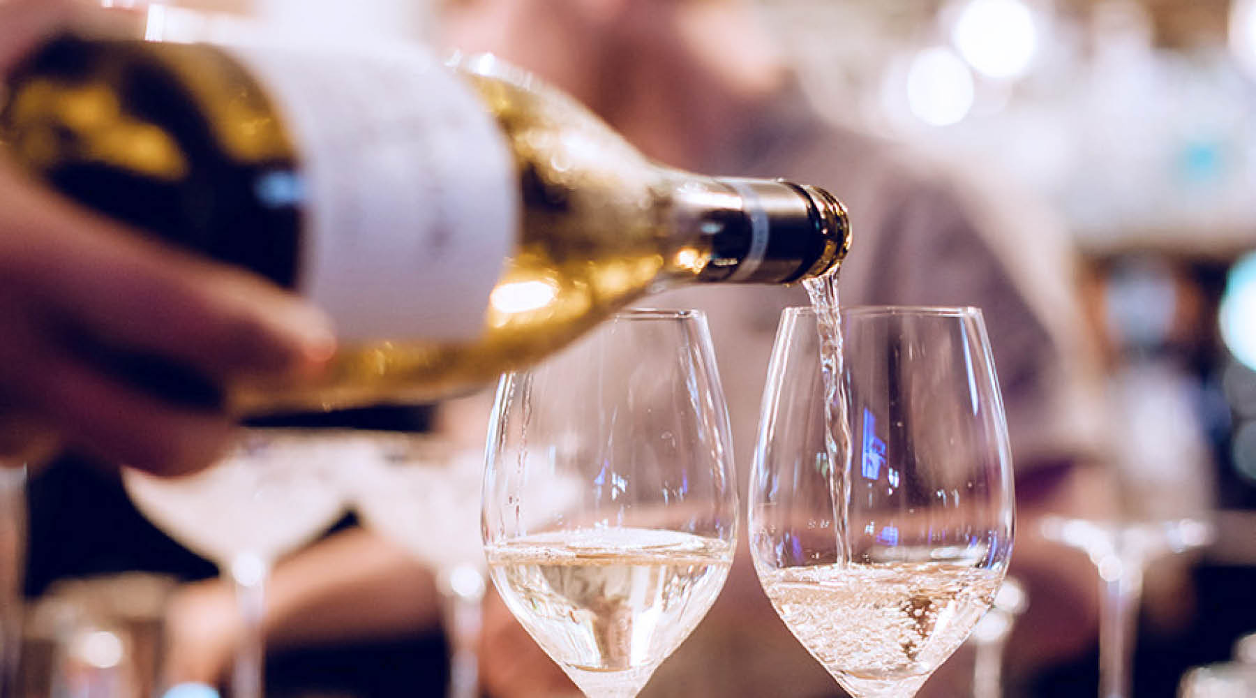 A close-up of a bottle of Jules Taylor white wine being poured into two clear wine glasses, with a blurred person and bar-like background visible.
