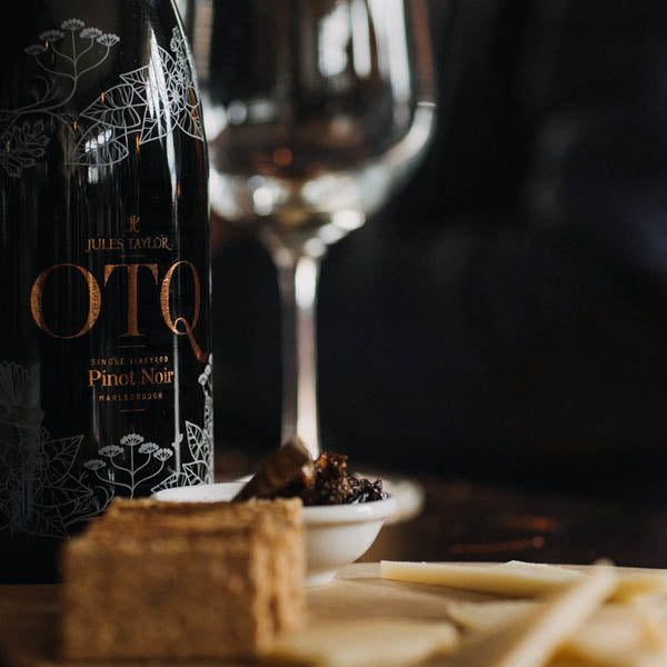A bottle of Jules Taylor On The Quiet Pinot Noir with a wine glass and snacks against a dark background.